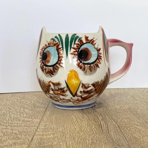 May include: A ceramic owl-shaped mug with a white base, featuring brown, blue, and yellow painted details. The mug has a pink handle and a blue stripe at the base. The owl's eyes are large and expressive.