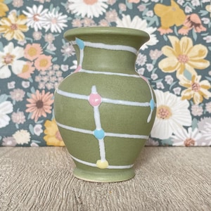 May include: A small, olive green ceramic vase with a rounded body and a flared rim. The vase is decorated with white horizontal stripes and vertical lines, accented with pink, blue, and yellow dots. The vase is approximately 13 cm tall.