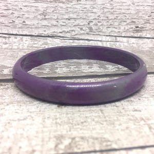 May include: A solid purple bangle. The bracelet is a deep shade of purple with a glossy finish. The bangle is circular and appears to be made of a smooth, solid material. The jewellery is displayed on a light-coloured wooden surface.