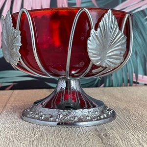 May include: A vintage red glass bowl with a silver metal frame and base. The bowl has a rounded shape and is adorned with silver leaf accents. The base is detailed with a decorative pattern, creating an elegant centrepiece.