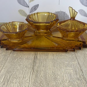 Könnte beinhalten: Bernsteinfarbenes Glasservierset mit einem rechteckigen Tablett und drei separaten Schalen. Das Set umfasst zwei kleine Schalen und eine Schale mit Deckel, alle mit geometrischen Art-Déco-Designs. Das Tablett hat ein Sonnenstrahlenmuster.