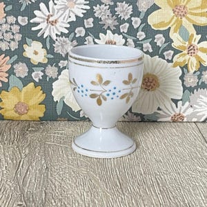 Könnte beinhalten: Weißer Eierbecher aus Keramik mit goldenem Rand und floralem Design. Der Eierbecher hat goldene Blätter und blaue Punkte. Der Sockel ist im Sockelstil gehalten. Der Hintergrund ist ein Blumenmuster.