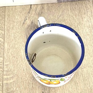 May include: A white enamel mug with a blue rim and a floral design featuring pears. The mug has a handle and shows signs of wear and tear.