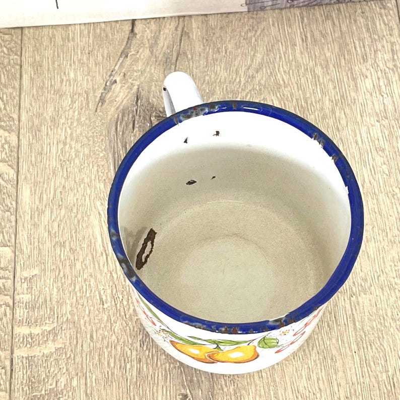 May include: A white enamel mug with a blue rim and a floral design featuring pears. The mug has a handle and shows signs of wear and tear.