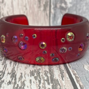 Könnte beinhalten: Ein breites, rotes Armreif-Armband mit schillernden Strasssteinen. Das Armband hat eine tiefrote Farbe und eine glänzende Oberfläche. Die Strasssteine sind in verschiedenen Größen und reflektieren mehrere Farben. Ein stilvolles Accessoire.