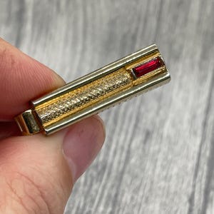 May include: A gold-coloured tie clip with a textured central section and a rectangular red gemstone. The clip has a classic design, suitable for formal wear. The gemstone adds a pop of colour to the accessory.