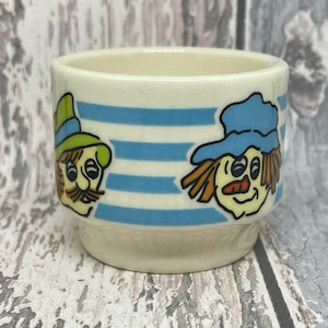 May include: A small, off-white ceramic cup with blue horizontal stripes. The cup features cartoon illustrations of two characters with hats and smiling faces. One character has a green hat and mustache, while the other has a blue hat and orange hair.