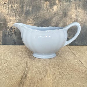 Könnte beinhalten: Eine hellblaue Keramik-Sauciere mit gewelltem Rand und gebogenem Griff. Die Sauciere steht auf einer Holzoberfläche. Die Sauciere ist ein klassisches Design zum Servieren von Saucen und Soßen.