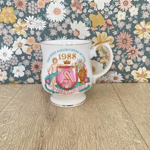 May include: A white ceramic mug with gold trim and handle. The mug features a pink crest with the letter "A" and the text "THE DUKE AND DUCHESS OF YORK 1988 TO CELEBRATE THE BIRTH OF THEIR FIRST CHILD".