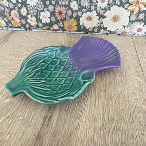 May include: A ceramic artichoke-shaped dish with a teal green base and a purple top. The dish has a textured surface with detailed leaf patterns. The background features a floral pattern on a light blue surface.