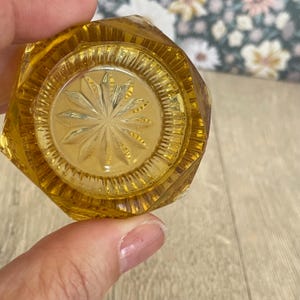 May include: A close-up of a vintage, amber-colored glass door knob. The knob has a faceted, octagonal shape with a detailed, starburst design in the center. The glass is clear, allowing light to pass through, and is held by a person's fingers.