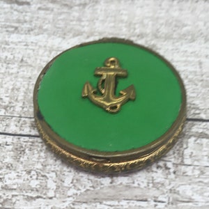 May include: A round, vintage compact with a bright green center and a gold-colored anchor emblem. The compact has a gold-colored, ornate border. The background is a light-colored, textured surface.