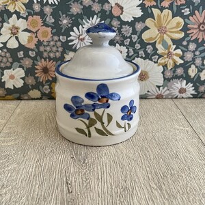 May include: A white ceramic sugar bowl with a matching lid, featuring hand-painted blue flowers with green stems and leaves. The bowl has a rounded shape with a blue rim accent and a small knob handle on top of the lid, showcasing a floral design.