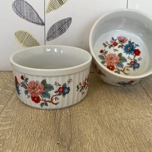 May include: Two white ceramic ramekins with a floral design. The ramekins have a ribbed exterior and a floral pattern of pink, red, and blue flowers on a white background. One ramekin is upright, and the other is tilted.