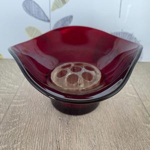 Könnte beinhalten: Eine tiefrote Glasschale mit einem transparenten Blumeneinsatz. Die Schale hat eine ausgestellte, asymmetrische Form und einen dunklen Rand. Der Blumenfrosch hat mehrere Löcher zum Anordnen von Stielen.