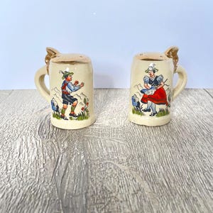 May include: A pair of ceramic salt and pepper shakers shaped like miniature beer steins. Each stein is beige with a handle and a decorative lid. One features a man in traditional German attire, the other a woman in a red skirt.
