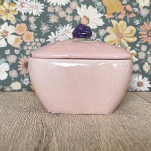 May include: A pink ceramic trinket box with a lid. The box has a textured surface and a purple berry-shaped knob on top. The background features a floral pattern.