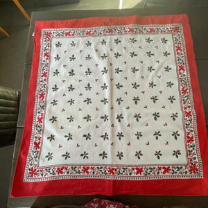 May include: A square tablecloth featuring a white centre with a repeating pattern of green holly leaves and red berries. The border consists of a decorative band of red poinsettia flowers and green holly leaves, finished with a solid red edge.