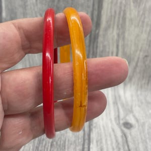 May include: Two thin, circular bangle bracelets. One bracelet is a solid, opaque red colour with a smooth, glossy finish. The second bracelet is a translucent, golden-orange colour with a smooth, glossy finish and visible internal air bubbles.