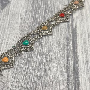 May include: Silver-toned bracelet featuring heart-shaped links. Each heart is adorned with a different colored enamel: orange, green, yellow, red, and pink. The bracelet is displayed on a gray wooden surface.