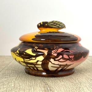May include: A ceramic jar with a lid, hand-painted with yellow, brown, and pink hues. The lid has a small, leaf-shaped handle. The jar is likely for storage or decorative purposes, with a unique, artistic design.