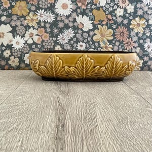 May include: A mustard-colored ceramic planter with a leaf design. The rectangular planter has a dark brown interior and a raised leaf pattern around the sides. The background features a floral pattern.