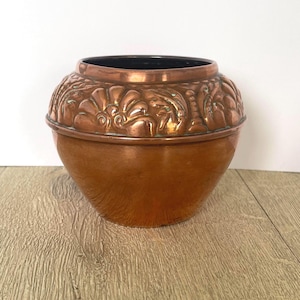 May include: A copper-coloured, round planter with a decorative floral pattern around the top. The planter has a dark interior and sits on a wooden surface. The planter is a vintage piece.