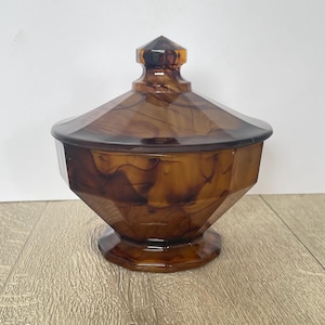 May include: A decorative brown lidded jar with a faceted design. The jar has a hexagonal base and a matching lid with a pointed finial. The translucent material has a marbled appearance.