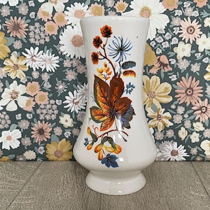 May include: A white ceramic vase with a flared rim and base, featuring a colorful botanical print on the front. The design displays detailed orange, brown, and blue leaves, berries, and small flowers arranged in a vertical, flowing pattern.