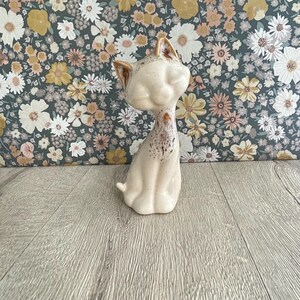 May include: A ceramic cat figurine with a cream-colored glaze and speckled brown accents on the ears, face, and chest. The cat is depicted in a seated position with its head tilted slightly to the side, featuring smooth, rounded features.