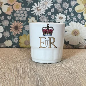 May include: A white ceramic mug featuring a graphic of a royal crown in red, blue, and gold above the gold-coloured letters E II R. The mug has a stepped, cylindrical base design with vertical ridges around the lower portion of the exterior.