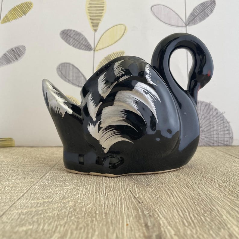 May include: A black ceramic swan figurine with silver painted details on the wings and body. The swan has a curved neck and a red accent on its beak. The figurine is a decorative item.
