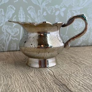 May include: A silver creamer with a scalloped rim and a curved handle. The creamer has etched floral designs around the body and sits on a small, round base. The creamer is displayed on a wooden surface.