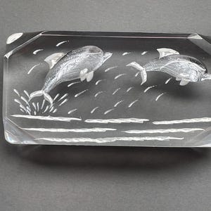 Puede incluir: Bloque de vidrio rectangular transparente con dos delfines saltando grabados. Los delfines están representados con detalles, sobre olas estilizadas. El bloque tiene bordes biselados y un acabado pulido, creando un efecto 3D.