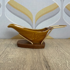 May include: A vintage brown ceramic gravy boat with a gold-coloured metal handle and stand. The word "SAUCE" is embossed on the side. It sits on a dark brown wooden base.