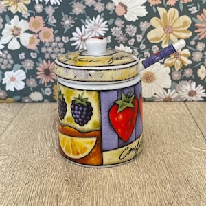 May include: A ceramic jam jar with a lid and matching spoon, featuring colorful illustrations of blackberries, a strawberry, and a lemon slice. The jar has the word Confiture written in black script on the side and lid, with a white background.