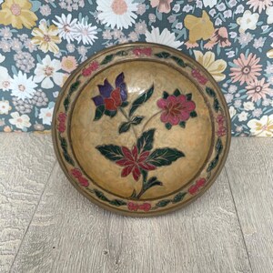 Könnte beinhalten: Eine runde, dekorative Schale mit floralem Design. Die Schale hat einen goldfarbenen Rand und eine beige Innenseite. Das florale Design zeigt rote, rosa und lila Blumen mit grünen Blättern.