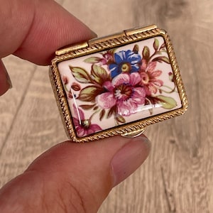May include: A small rectangular metal pill box with a hinged lid featuring a glossy ceramic inlay. The inlay displays a colourful floral pattern with pink, blue, and green flowers on a white background. The metal frame has a textured rope-like edge.