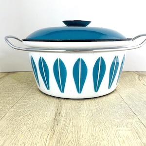 May include: A white enamel pot with a teal lid and handles. The pot has a teal leaf pattern around the exterior. The lid has a black trim and a small, dark teal knob. The pot is on a wooden surface.