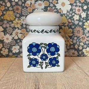 May include: A white ceramic jar with a lid, decorated with blue floral patterns. The jar has a square shape with rounded corners and a green leaf design around the top. The lid is a rounded shape.