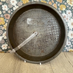 May include: A circular, vintage metal cake tin with a textured base and a handle. The tin has a dark, aged appearance, showing signs of use. The interior has the text "DURVENE" and "REGD TRADE MARK". Made in England.