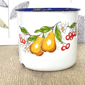 May include: White enamel mug with a blue rim and a floral design featuring pears, cherries, and white flowers.