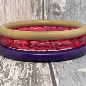 May include: Three stacked bangles in beige, pink, and purple. The top bangle is a solid beige colour, the middle one is a transparent pink with a textured pattern, and the bottom one is a solid purple. The bangles are stacked on top of each other.