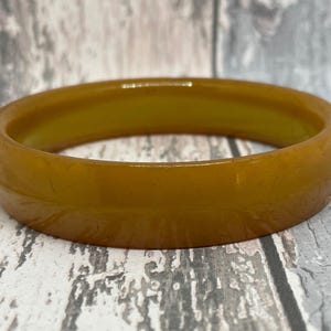 May include: A close-up of a thick, amber-coloured bangle bracelet. The bracelet has a smooth, glossy finish and a slightly translucent appearance. The colour is a warm, golden hue, with subtle variations in tone. The bracelet is a simple, yet elegant accessory.