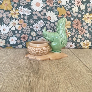 May include: A ceramic figurine featuring a light green squirrel positioned next to a small, round, tan-colored bowl with a textured, bumpy exterior. The squirrel has a curved, bushy tail and is attached to a smooth, flat, tan-colored base.