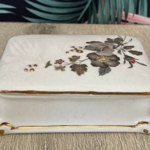 May include: A rectangular white ceramic jewellery box with a floral design on the lid. The flowers are grey and brown with gold accents. The box has a gold trim and sits on small feet.