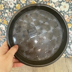 May include: A round, dark metal baking pan with a starburst pattern on the bottom. The pan has a raised edge and is being held by a hand. The text "MADE IN ENGLAND" and "OVENEX" are visible.