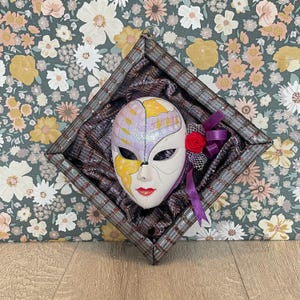 May include: A decorative mask with a half-face design, featuring purple and yellow accents, red lips, and a white base. The mask is set within a plaid-patterned frame, adorned with a red rose and purple ribbon.