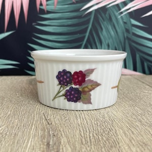 May include: A white ceramic ramekin with vertical ribbed detailing. The ramekin features a colorful illustration of blackberries and raspberries with green leaves. The ramekin is sitting on a wooden surface.