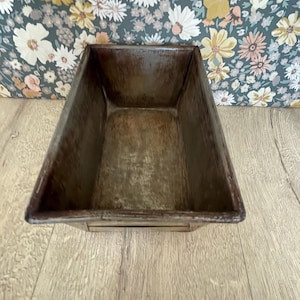 May include: A rectangular, brown metal container with a distressed finish. The container has a slightly tapered shape and appears to be empty. The background features a floral pattern on a dark background.